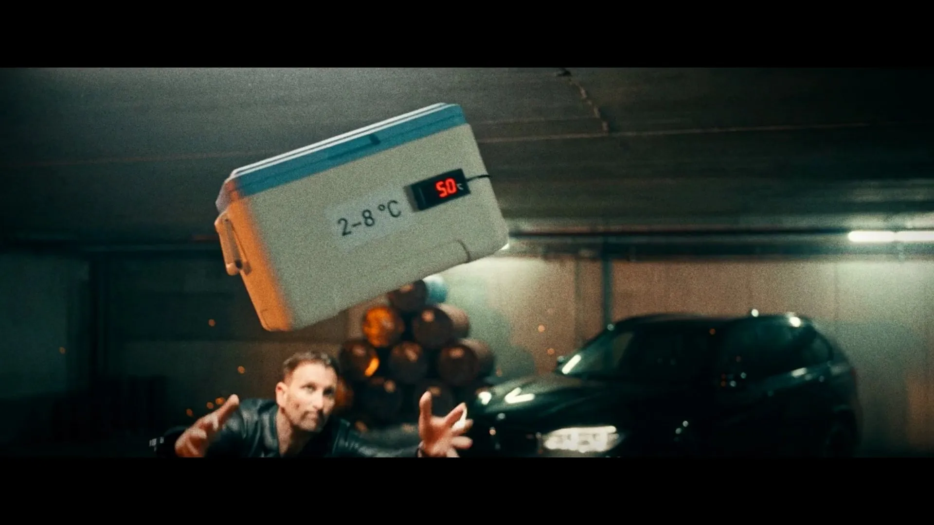 Standbild aus dem Grippe-Impfung-Trailer – in einer nächtlichen Tiefgarage fliegt eine weiße Kühlbox mit der Aufschrift „2–8 °C“ durch die Luft, darunter streckt ein Mann die Arme nach ihr aus
